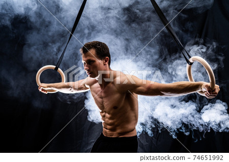 Muscular build man doing calisthenics on gymnastics rings indoor on black, smoked background. concept of motivation, desire and passion 74651992