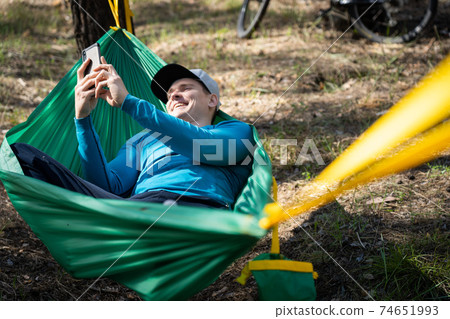Young happy man using mobile phone in hat relaxing outside in hammock in forest. 74651993