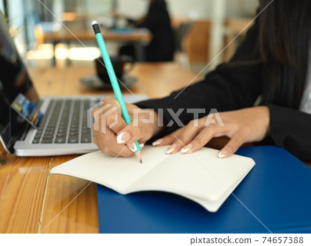 Businesswoman hand with pencil writing on notebook while working with laptop Businesswoman hand with pencil writing on notebook while working with laptop 74657388