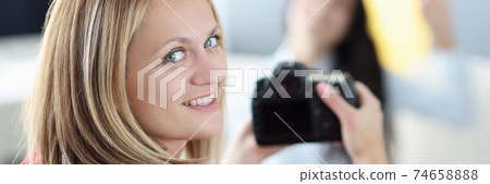 Young woman photographer holding camera in her hands with brunette in background at home Young woman photographer holding camera in her hands with brunette in background at home 74658888