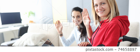 Young disabled woman in wheelchair with helper is sitting by laptop screen and waving hello 74658897