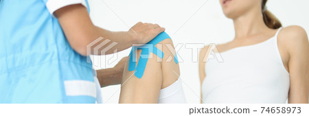 Traumatologist doctor supports knee with blue tape to young woman patient in clinic closeup Traumatologist doctor supports knee with blue tape to young woman patient in clinic closeup 74658973