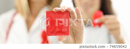 Two female doctors are holding condom and a red ribbon. 74659042