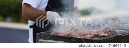 Man in black apron turn grilled meat with spatula om street. 74664640