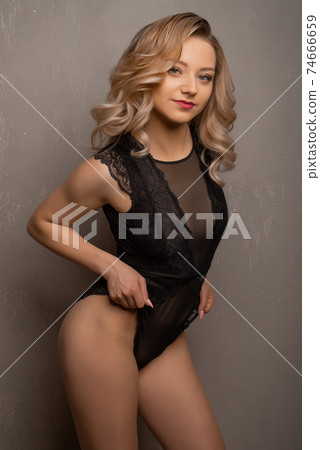 Beautiful young blonde woman posing in black lace bodysuit in the studio near the wall Beautiful young blonde woman posing in black lace bodysuit in the studio near the wall 74666659