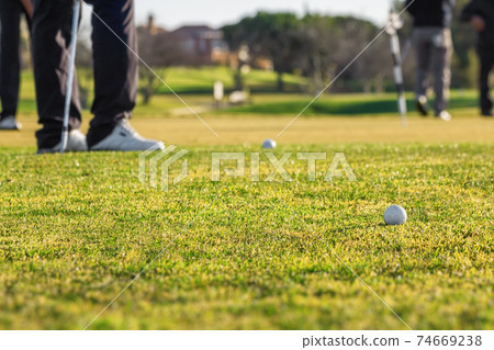 Golden hour shot of golf course with focus on a ball on the field. Legs of golfers can also be seen on the side. Golden hour shot of golf course with focus on a ball on the field. Legs of golfers can also be seen on the side. 74669238