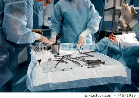 close up surgeons hand taking scissors, forceps and surgical instruments on table for operation with colleagues performing work in operation room at hospital, emergency case, surgery, medical concept 74671919
