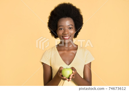 Beautiful african girl with perfect smile holding fresh green apple in hand isolated on bright yellow background. Healthy lifestyle, natural beauty, skincare, veganism, health care concept. 74673612