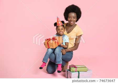 Child's birthday, holiday celebration concept. Cute happy little African girl in party hat, holding yellow present box, while sitting on her mommy's legs over pink studio background Child's birthday, holiday celebration concept. Cute happy little African girl in party hat, holding yellow present box, while sitting on her mommy's legs over pink studio background 74673647