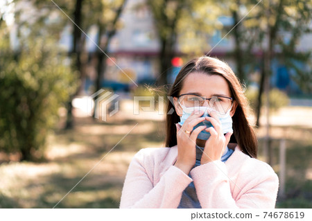care, infants, spring, coronavirus and quarantine concept - Young cute long haired woman European Caucasian Slavic appearance put on blue medical protective mask in midday sunlight backlight in park. 74678619