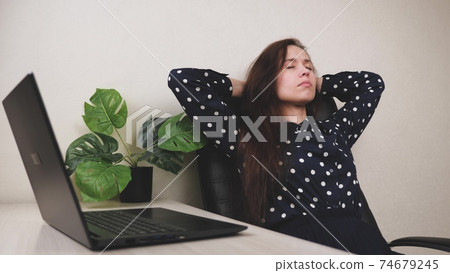 A serene attractive young woman is resting on an armchair, at a table with a laptop at work, deeply breathing fresh air, holding hands behind her head, a healthy calm lady is relaxing on a comfortable 74679245