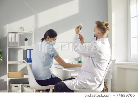 Male nurse drawing antiviral vaccine out of vial before giving shot to young woman 74689700
