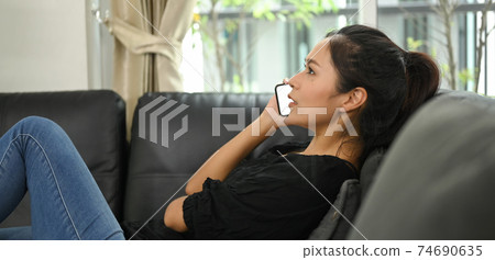 Young Asian woman is talking on a mobile phone while lying on the black leather sofa. Young Asian woman is talking on a mobile phone while lying on the black leather sofa. 74690635