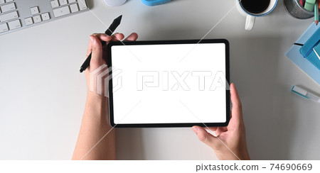 Hands are holding a white blank screen computer tablet and stylus pen over a white working desk. 74690669