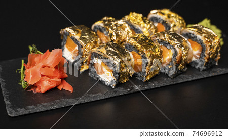 Custom sushi roll with nori, cuttlefish ink, fresh salmon, cream cheese, pepper pumpkin, eel, unagi sauce and edible gold leaf with red ginger, wasabi on a square plate on black table and background. 74696912