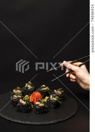 Hand with chopsticks wants to take custom sushi roll with black rice, crab meat, avocado, smoked salmon mousse, oar caviar, masago, shrimp cocktail, edible gold leaf, ginger, wasabi on black table. 74696914