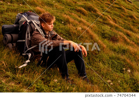 A young tourist sits on a background of mountains with a backpack, relaxing and admiring the views of the Carpathians in Ukraine. 74704344