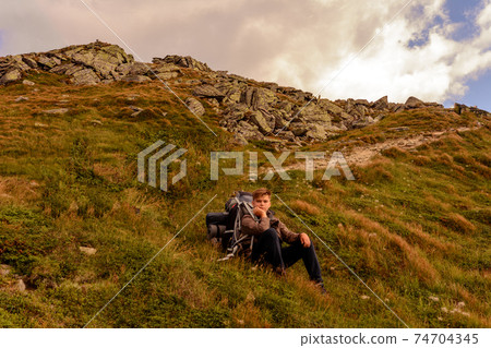 A young tourist sits on a background of mountains with a backpack, relaxing and admiring the views of the Carpathians in Ukraine. A young tourist sits on a background of mountains with a backpack, relaxing and admiring the views of the Carpathians in Ukraine. 74704345