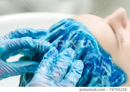 Hairdresser in protective gloves washes client's blue hair with shampoo for washing after process of coloring hair in sapphire color Hairdresser in protective gloves washes client's blue hair with shampoo for washing after process of coloring hair in sapphire color 74705226