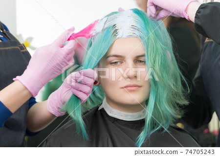 Process of bleaching hair roots in hair salon. Two hairdressers in glove use pink brush while applying paint to young adult customer with emerald hair color Process of bleaching hair roots in hair salon. Two hairdressers in glove use pink brush while applying paint to young adult customer with emerald hair color 74705243