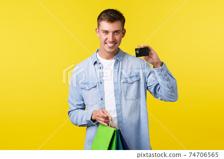 Shopping, leisure and discounts concept. Smiling handsome young man buying new clothes, holding bags and showing credit card with satisfied expression, paying with saved money, yellow background Shopping, leisure and discounts concept. Smiling handsome young man buying new clothes, holding bags and showing credit card with satisfied expression, paying with saved money, yellow background 74706565