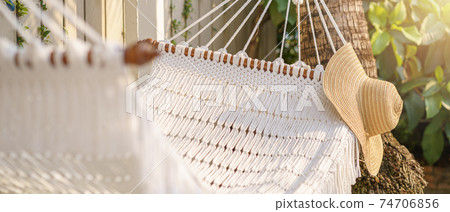 Straw hat hanging on white hammock outdoor. Relaxing summer vacation concept. Selective focus. Straw hat hanging on white hammock outdoor. Relaxing summer vacation concept. Selective focus. 74706856