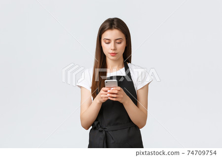 Grocery store employees, small business and coffee shops concept. Serious young female barista, waitress on break in black apron using smartphone, looking focused at screen 74709754