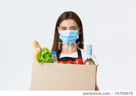 Covid-19 social distancing, delivery and grocery shopping during coronavirus concept. Young pleasant saleswoman, cashier in medical mask handling over groceries to customer 74709879