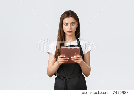 Grocery store employees, small business and coffee shops concept. Young female cafe worker in black apron manage website or social media. Barista holding digital tablet and smiling camera Grocery store employees, small business and coffee shops concept. Young female cafe worker in black apron manage website or social media. Barista holding digital tablet and smiling camera 74709881