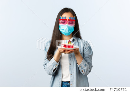 Social distancing lifestyle, covid-19 pandemic, celebrating holidays during coronavirus concept. Hopeful cute asian birthday girl in medical mask and party glasses, making wish over b-day cake 74710751