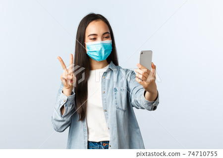 Social distancing lifestyle, covid-19 pandemic and people emotions concept. Friendly cheerful asian woman in medical mask taking selfie for social media, make peace sign at smartphone camera Social distancing lifestyle, covid-19 pandemic and people emotions concept. Friendly cheerful asian woman in medical mask taking selfie for social media, make peace sign at smartphone camera 74710755