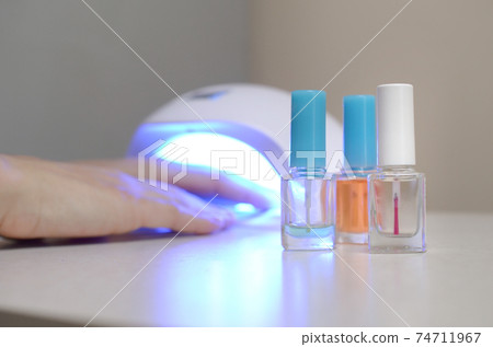 Three colorless transparent nail polish bottles in background of female hands in gel uv led nail white lamp for drying manicure 74711967