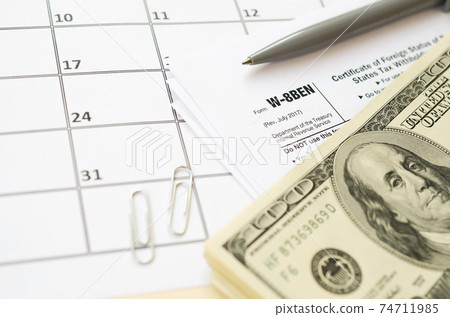 IRS Form W-8BEN Certificate of foreign status of beneficial owner for United States tax withholding and reporting for individuals blank lies with pen and hundred dollar bills on calendar page IRS Form W-8BEN Certificate of foreign status of beneficial owner for United States tax withholding and reporting for individuals blank lies with pen and hundred dollar bills on calendar page 74711985
