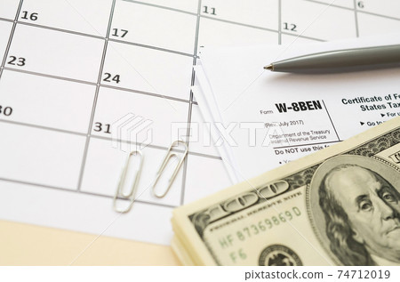 IRS Form W-8BEN Certificate of foreign status of beneficial owner for United States tax withholding and reporting for individuals blank lies with pen and hundred dollar bills on calendar page 74712019