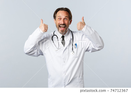 Covid-19, coronavirus outbreak, healthcare workers and pandemic concept. Excited upbeat male doctor in white coat thumbs-up in approval. Surgeon agree or like idea, recommend promo 74712367