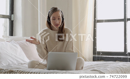 Smiling woman wearing headset looking at laptop, sitting on her bed with laptop, making video call 74722099