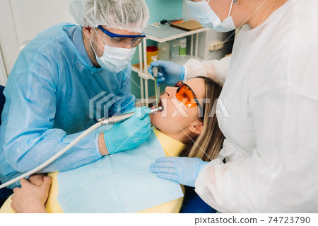 A male dentist with dental tools drills the teeth of a patient with an assistant. The concept of medicine, dentistry and healthcare 74723790