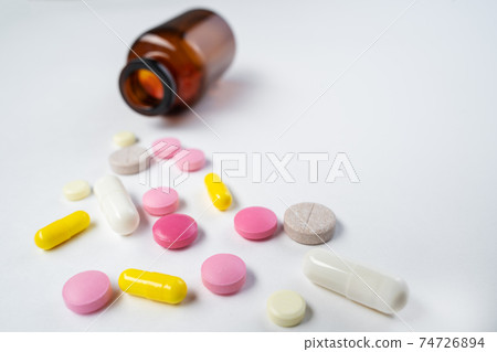 Multicolor pills and tablets spilling out from glass pill bottle on white. Multicolor pills and tablets spilling out from glass pill bottle on white. 74726894
