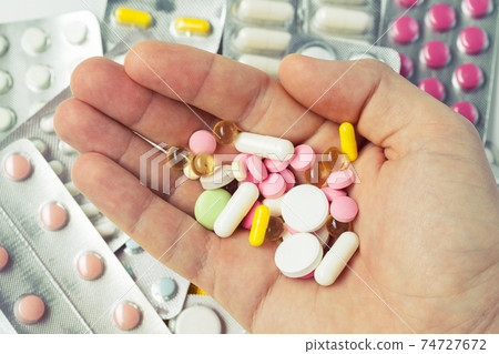 A handful of various pills in a man's hands. Medications against the COVID-19 and other conditions and diseases on the grey background. A handful of various pills in a man's hands. Medications against the COVID-19 and other conditions and diseases on the grey background. 74727672