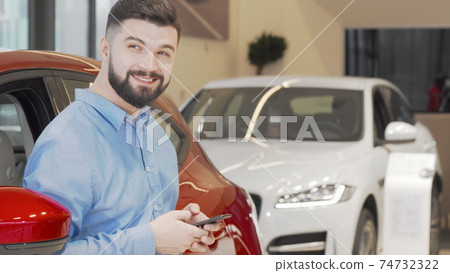 Happy man texting on a smart phone while shopping for new car at the dealership 74732322