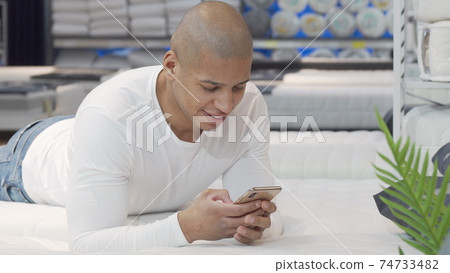 Young African man using smart phone while trying new bed at furniture store 74733482
