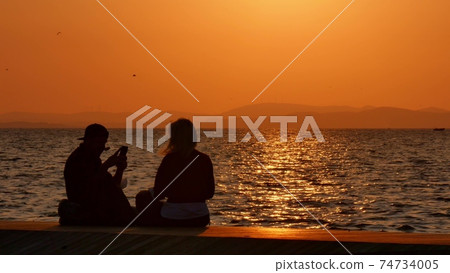 Silhouette of a young couple taking a selfie at sunset, with a sea and sunset in the background. 74734005