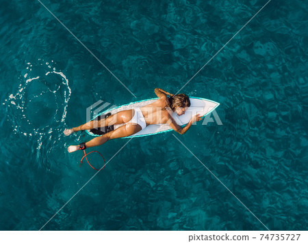 Attractive surfer woman relaxing on surfboard in blue ocean. Aerial view with surf girl Attractive surfer woman relaxing on surfboard in blue ocean. Aerial view with surf girl 74735727