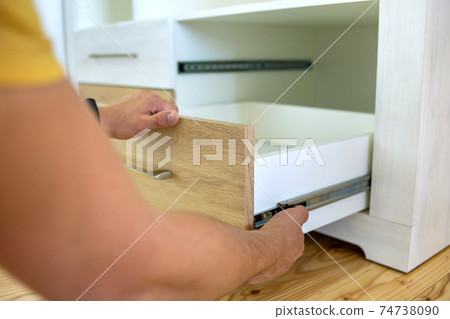 Close up of carpenter hands installing wooden drawer on sliding skids in contemporary cupboard cabinet. 74738090