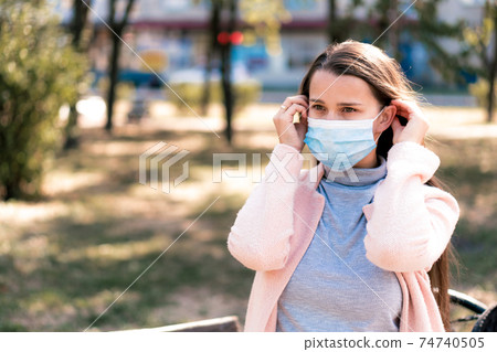 care, infants, spring, coronavirus and quarantine concept - Young cute long haired woman European Caucasian Slavic appearance put on blue medical protective mask in midday sunlight backlight in park. 74740505