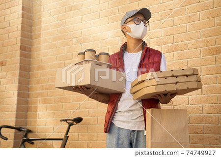 Male courier wearing face protective mask holding various packages with food, pizza boxes and four coffee cups while standing against brick wall 74749927