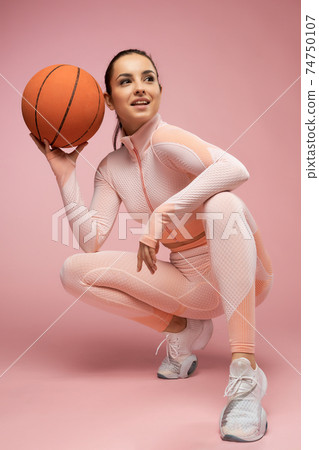 Joyful young woman with basketball ball crouching down in studio 74750107