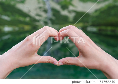 Womans making heart shape with her hands against the mountains near to the lake. Womans making heart shape with her hands against the mountains near to the lake. 74762210