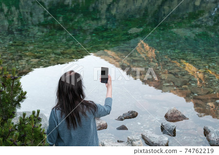 Travel bloger young woman standing on the lake shore and making photo with her smartphone.  74762219