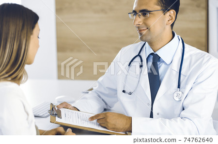 Doctor and patient discussing medical exam results while sitting at the desk in clinic. Male physician using clipboard for filling up medication history record of young woman. Data in medicine Doctor and patient discussing medical exam results while sitting at the desk in clinic. Male physician using clipboard for filling up medication history record of young woman. Data in medicine 74762640